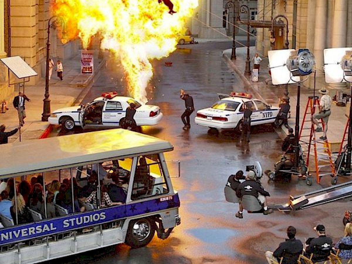 This image shows a movie set with an explosion, police cars, crew members filming, and a tram labeled 