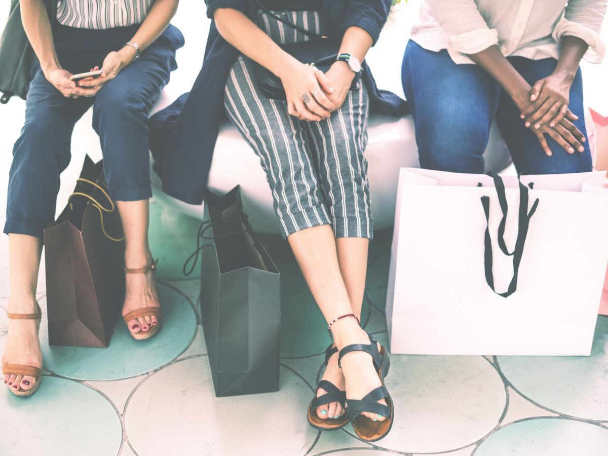 Three seated individuals holding shopping bags. They appear to be resting after a shopping trip. The focus is on their legs and bags.