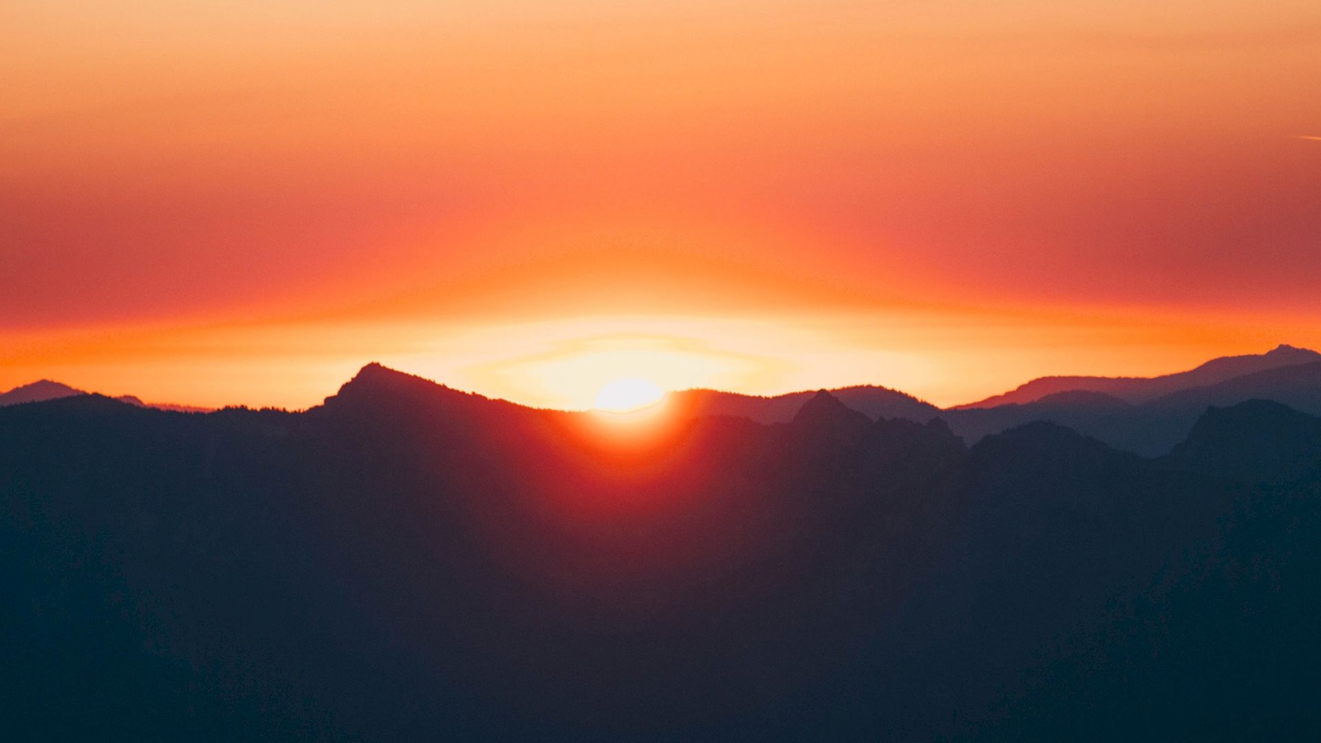 This image shows a stunning sunset behind the silhouette of distant mountains, with a vibrant sky transitioning from orange to purple hues.