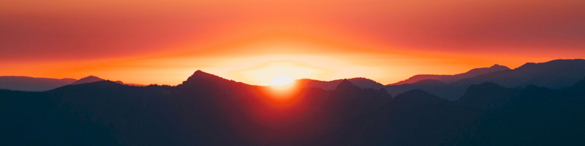 This image shows a stunning sunset behind the silhouette of distant mountains, with a vibrant sky transitioning from orange to purple hues.