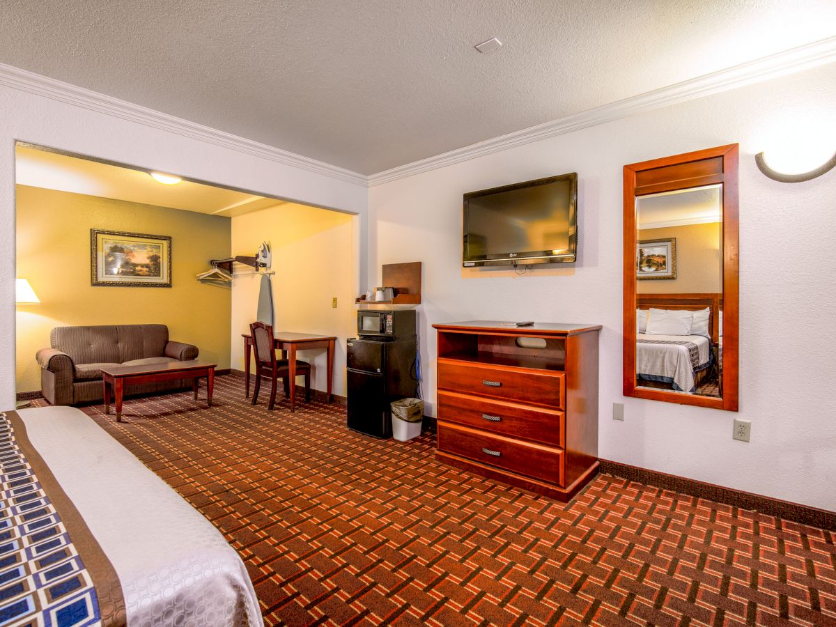 This image shows a hotel room with a bed, a TV on a dresser, a mini-fridge, a mirror, and a seating area with a couch and table.