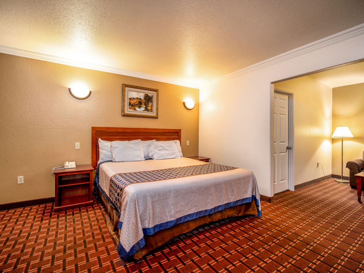 The image shows a neatly arranged hotel room with a bed, bedside tables, lamps, and a sitting area with a chair and floor lamp.