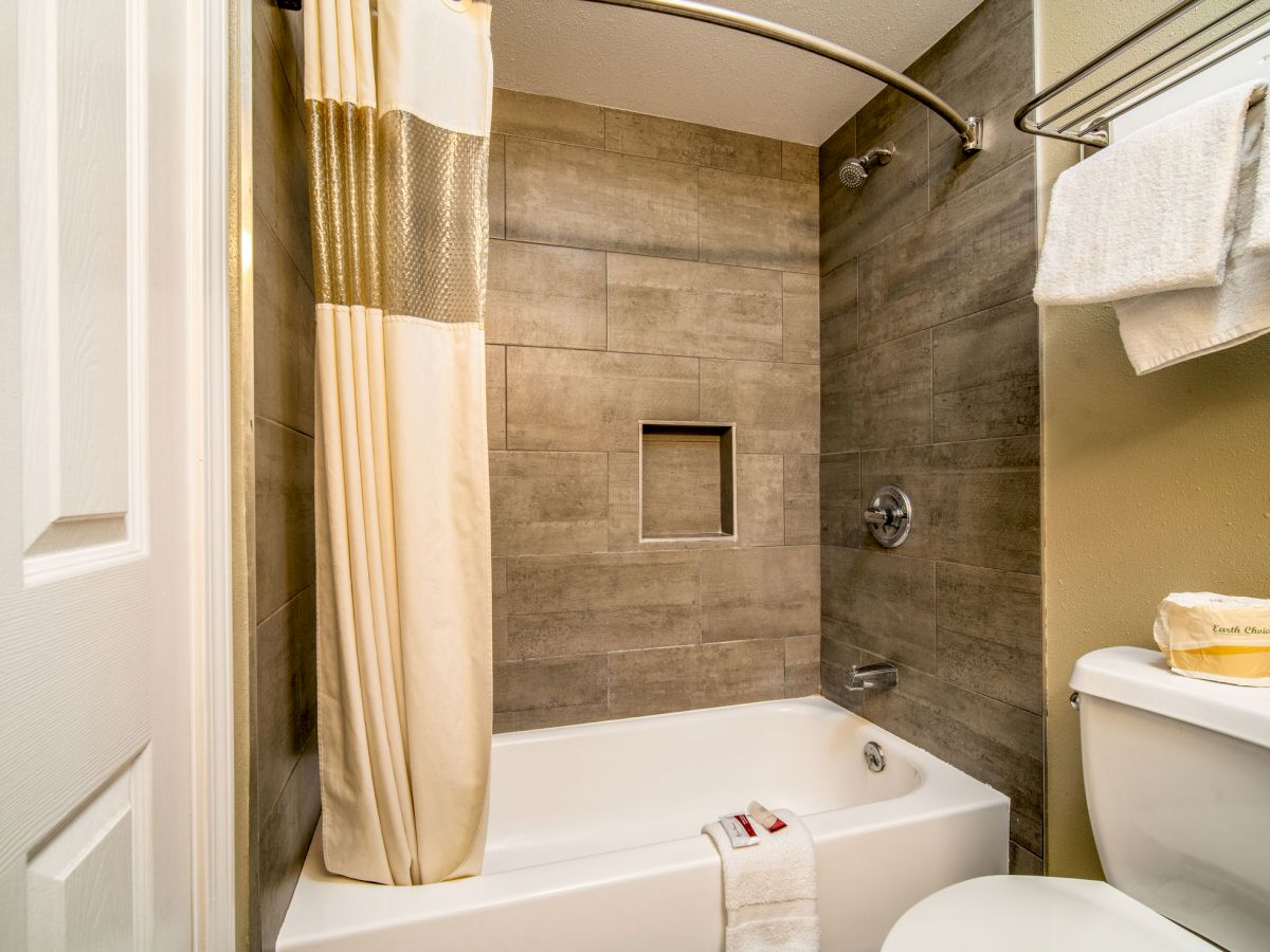 A clean bathroom with a shower and bathtub, beige tiles, a curtain, a towel on the tub edge, and another on a rack above the toilet, ending the sentence.