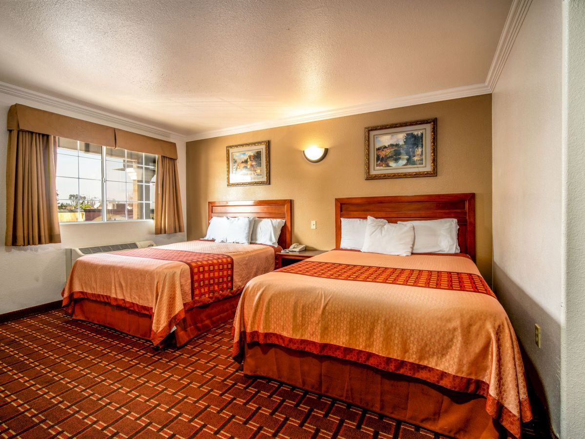 A hotel room features two double beds with orange bedspreads, wall decor, a window with curtains, and patterned carpet flooring, ending the sentence.