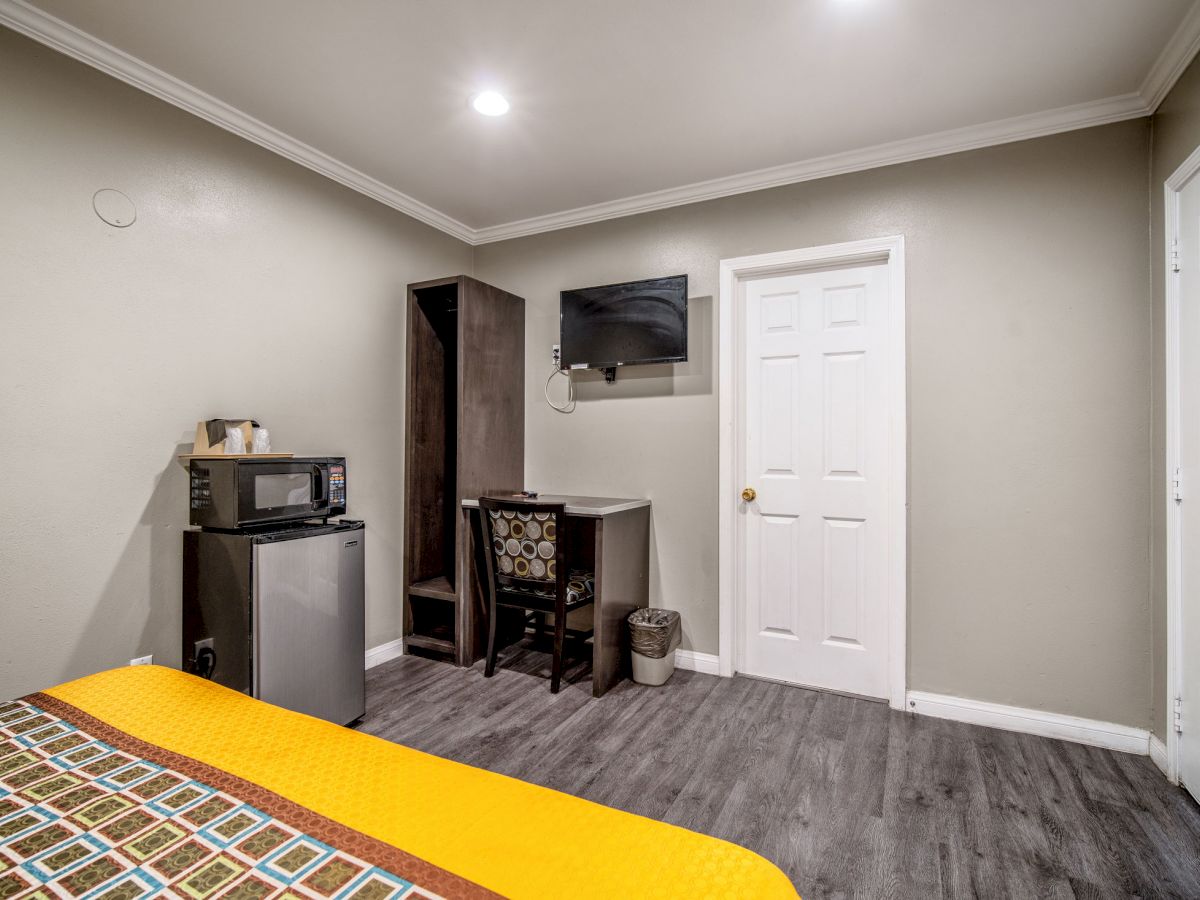 A small room with a bed, microwave, mini-fridge, desk, chair, mounted TV, and door; lit by ceiling lights.