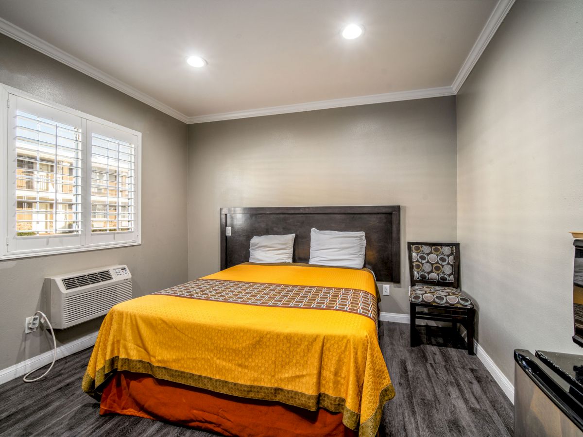 A small bedroom with a yellow bedspread, pillows, a window, an air conditioning unit, and a chair next to the bed in a beige-painted room.