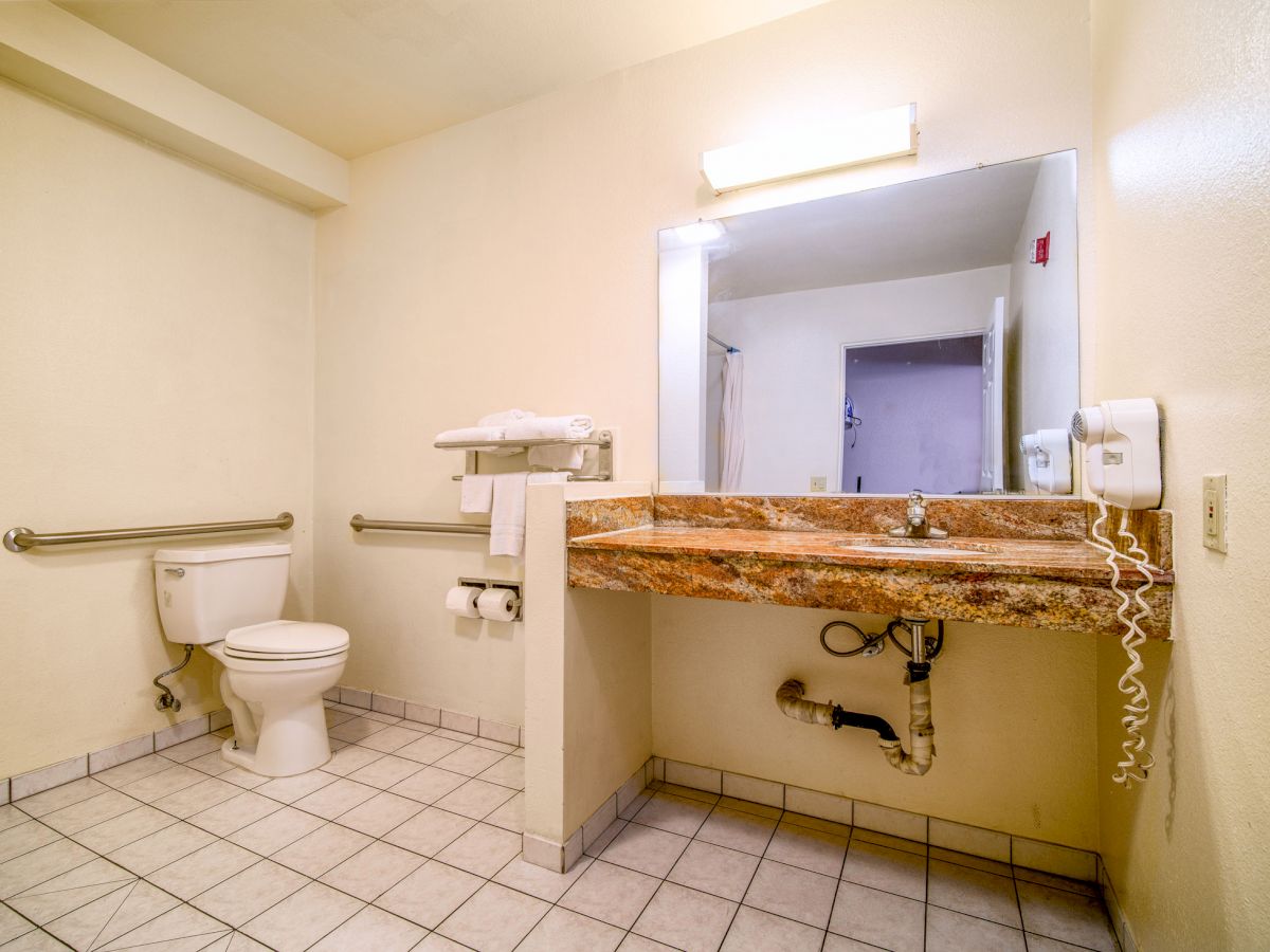 A bathroom with a toilet, a large mirror above a granite countertop sink, towel rack, hairdryer, and handrails on beige tiled floor ending the sentence.