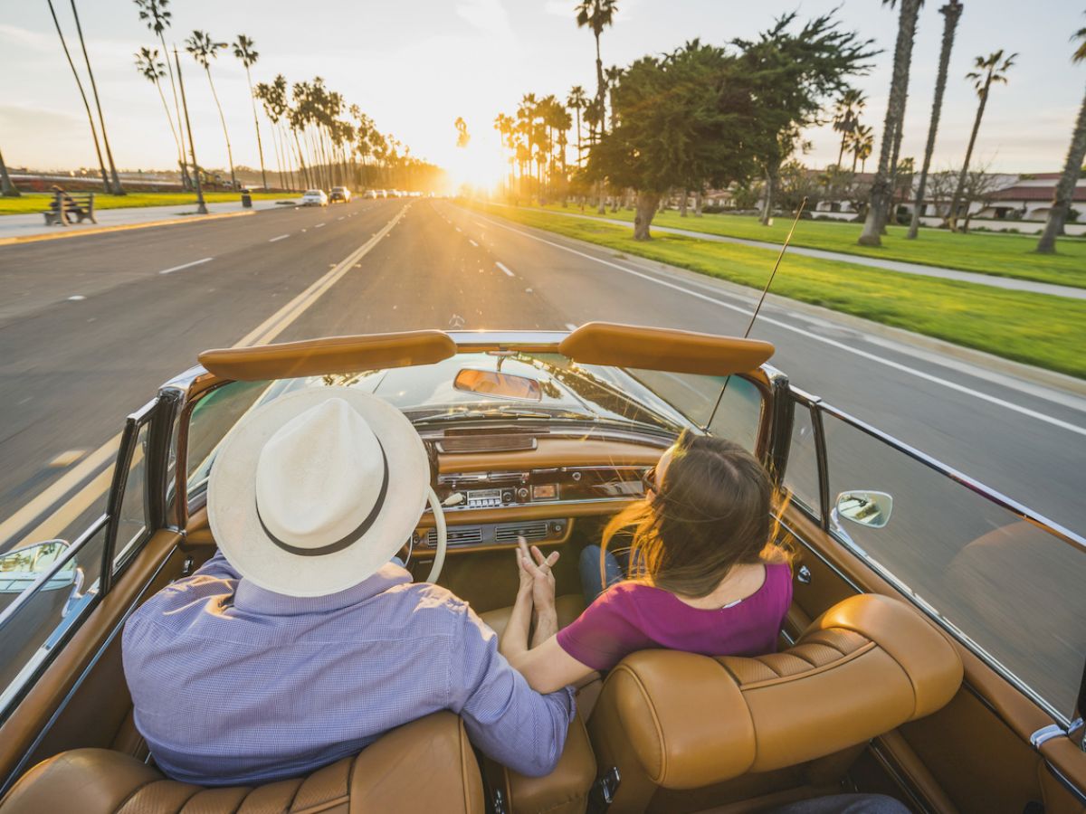 A couple is driving in a convertible on a palm-lined road at sunset, with the man wearing a hat and the woman leaning on his shoulder.