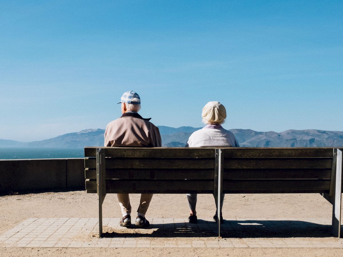 Two elderly individuals sit on a bench overlooking a serene landscape with mountains and a blue sky in the background, enjoying the view.