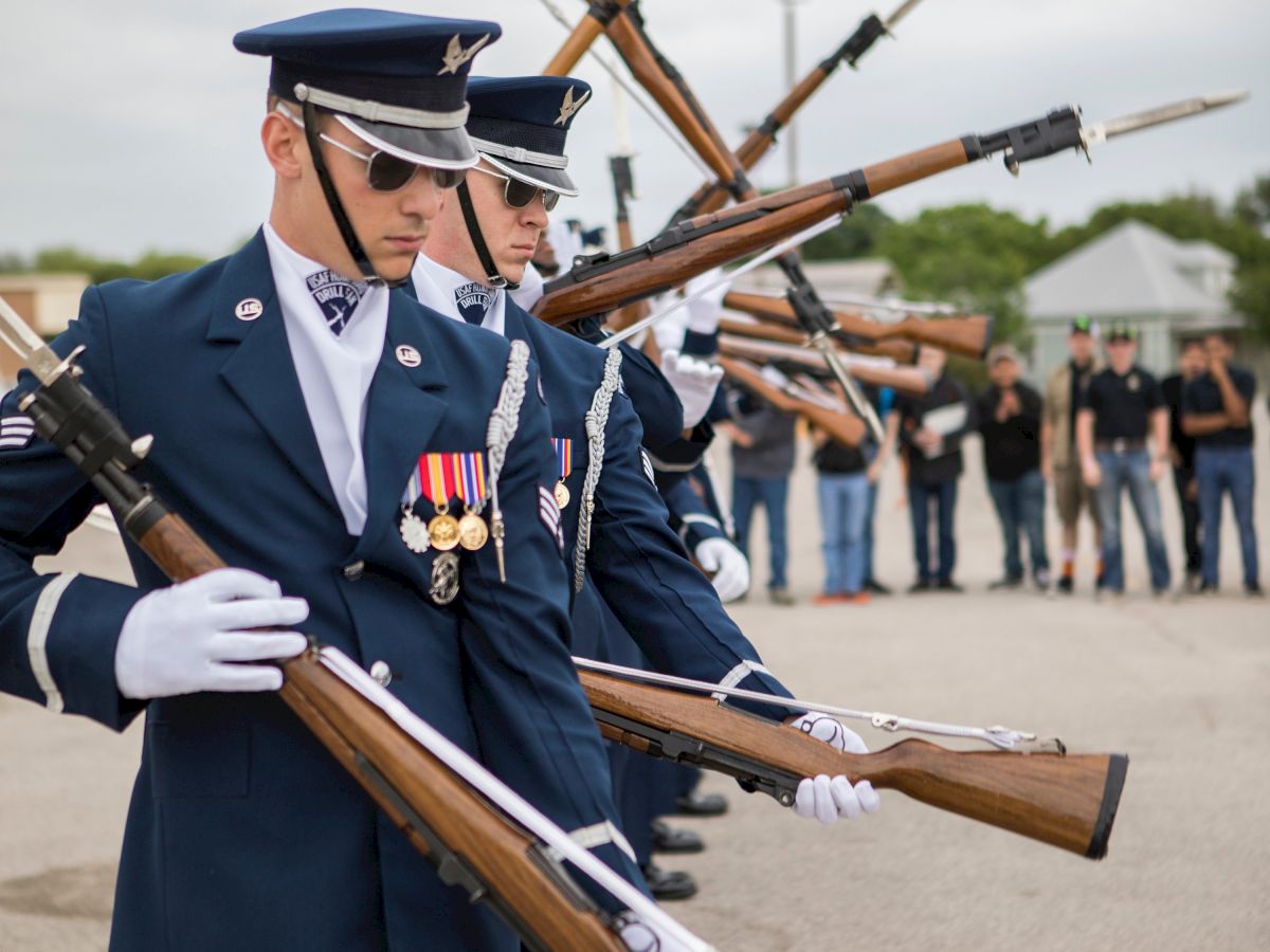 Several uniformed individuals are performing a synchronized rifle drill outdoors, while onlookers watch in the background.