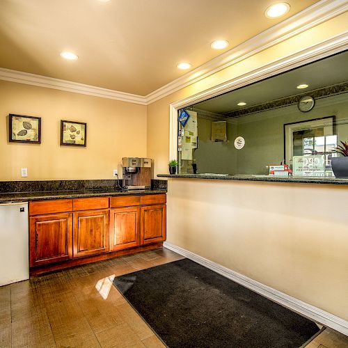 The image shows a reception area with a counter, wooden cabinets, a small refrigerator, a coffee maker, framed pictures on the wall, and overhead lighting.