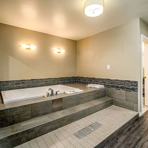 A modern bathroom with a sunken tub, dual wall lights, tiled walls and steps, and a dark wood floor, illuminated by a ceiling light.