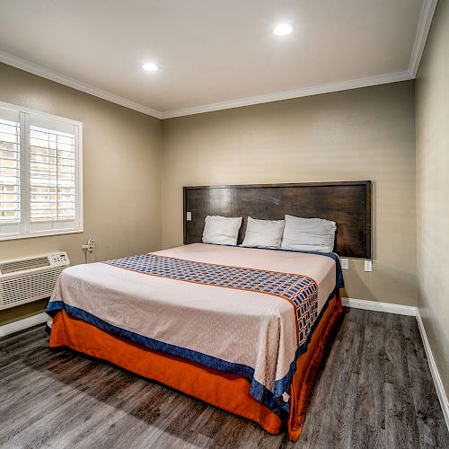 A minimalist bedroom with a large bed, simple bedding, wooden flooring, a window, and an air conditioner on the wall underneath the window.