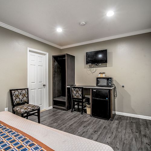 The image shows a hotel room with a bed, two chairs, a TV mounted on the wall, a microwave, a mini-fridge, and a small desk.