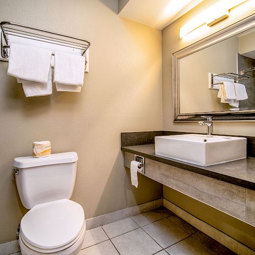 A modern bathroom with a toilet, a wall-mounted towel rack, a large mirror, and a square sink on a counter with a light above.