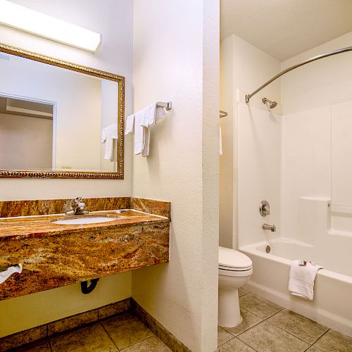 A bathroom with a marble counter, large mirror, sink, towel rack, toilet, and a bathtub with a curved shower curtain rod, all well-lit.