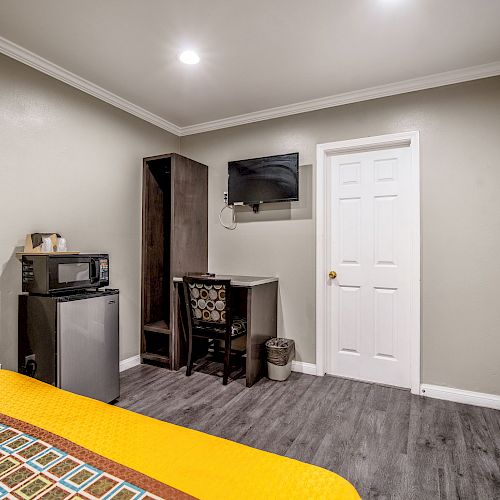 A hotel room with a bed, desk, chair, wall-mounted TV, microwave, mini-fridge, and door, and hardwood floor.