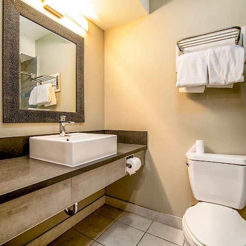 A bathroom with a sink, a mirror, a towel rack with folded towels, a toilet, and toilet paper; bright lighting above the mirror.