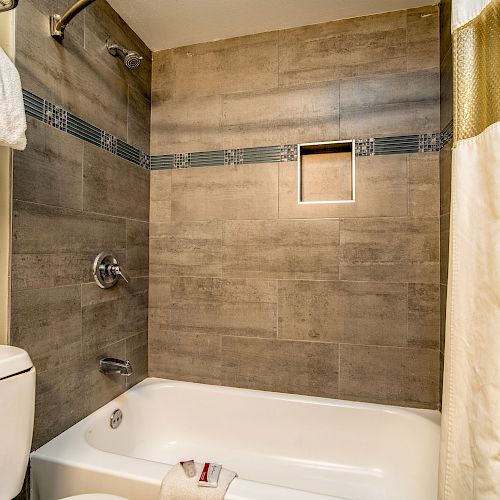 The image shows a modern bathroom with a bathtub, tiled walls, a showerhead, a toilet, and towels, along with a curtain partially drawn.