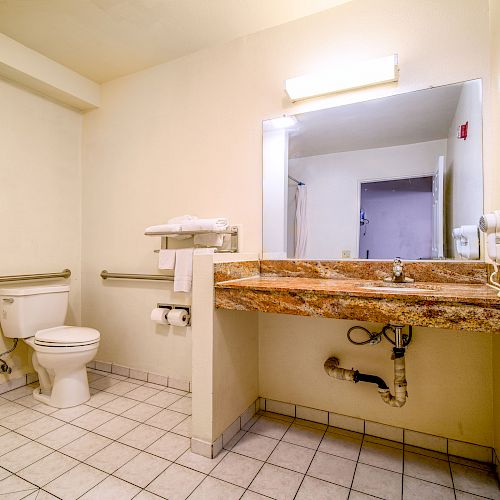 The image shows a bathroom with a toilet, a marble-topped sink, a mirror, a wall-mounted hair dryer, and white tile flooring.