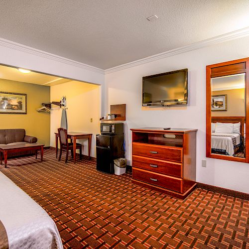 A cozy hotel room with a bed, TV, dresser, mirror, couch, table, chairs, and hangers. The room has patterned carpet and warm lighting.