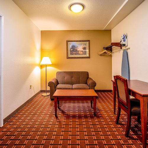 The image shows a small room with a sofa, coffee table, lamp, wall painting, desk with chair, ironing board, and a closet shelf with hangers.