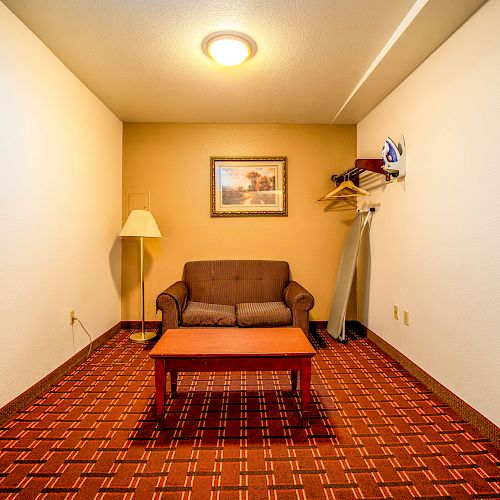 A small room with a patterned carpet, a brown loveseat, a wooden coffee table, a standing lamp, a wall-mounted picture, and a coat rack.