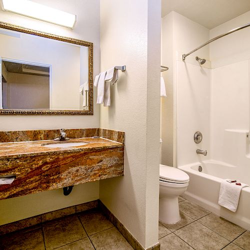 A well-lit bathroom with a large mirror above a granite countertop, toilet next to a bathtub/shower combo, and towels on the wall.