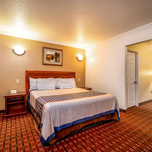 The image shows a neatly furnished hotel room with a bed, bedside tables, a lamp, and a painting on the wall, creating a cozy atmosphere.