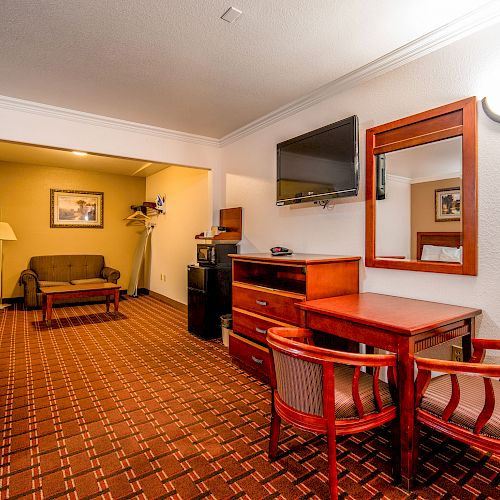 This image shows a hotel room with a bed, two chairs at a table, a TV on the wall, and a small lounge area with a sofa and lamp.