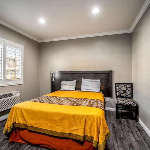 A neatly arranged bedroom with a queen bed, yellow bedspread, window, chair, air conditioner, and hardwood floor.