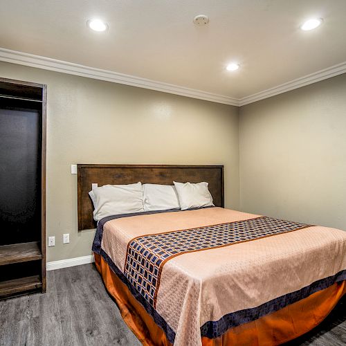 The image shows a simple, well-lit bedroom with a bed, headboard, three pillows, a patterned bed cover, and an empty open closet on the left side.