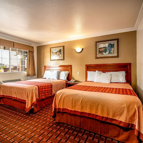 The image shows a hotel room with two double beds, a window with curtains, framed artworks on the wall, and a patterned carpet.