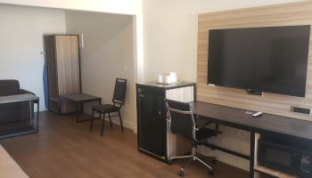 Small office room with desk, rolling chair, wall screen/TV, cabinets, and a door with a bench and chair.