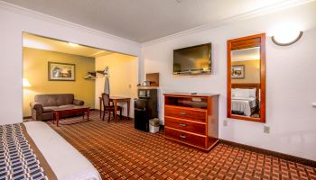 A well-lit hotel room with a bed, TV, dresser, mini-fridge, and a seating area including a sofa and a table, neatly arranged.