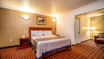 This image shows a hotel room with a neatly made bed, nightstands, wall art, and a brightly lit open space leading to a sitting area with a lamp.