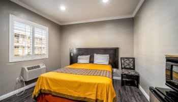 A small, well-lit bedroom with a bed, yellow bedspread, two pillows, a nightstand, chair, air conditioner, and a window with blinds.