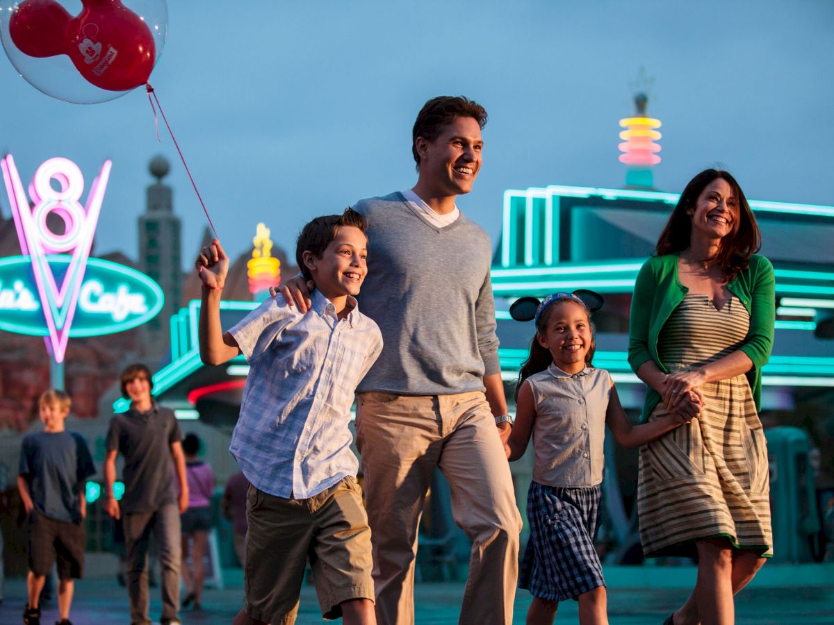 A family happily walks in a theme park with neon lights in the background, and the son holds a red balloon.