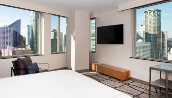 A modern hotel room with large windows, a TV, a chair, a small table, and a bed, offering a cityscape view from both sides of the room.