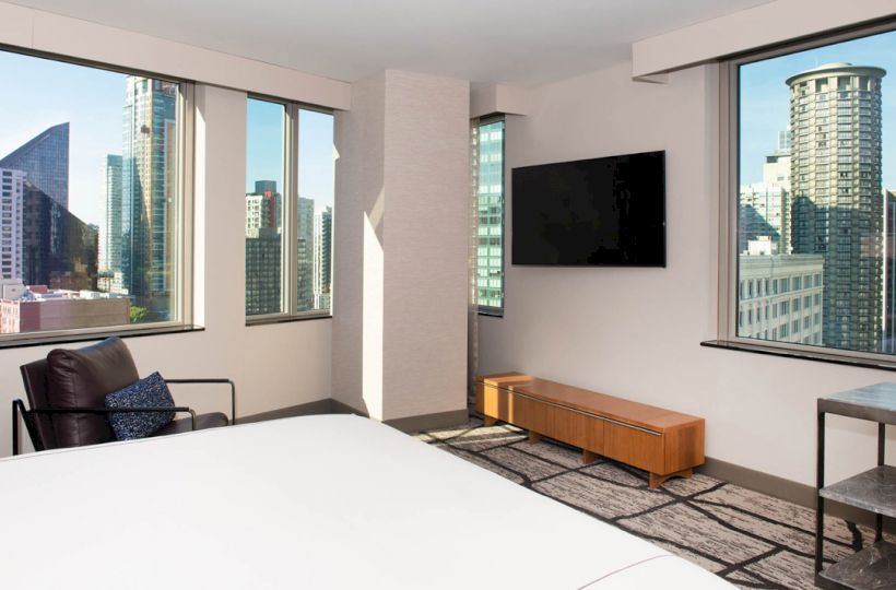 A modern hotel room with large windows, a TV, a chair, a small table, and a bed, offering a cityscape view from both sides of the room.