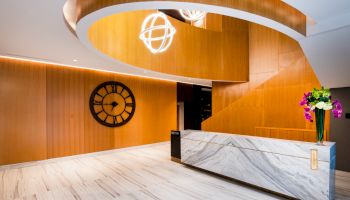 A modern lobby features a marble reception desk, wooden details, a large wall clock, modern lighting fixtures, and a flower arrangement, ending the sentence.