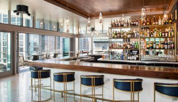 A modern bar with a wooden counter, blue and gold bar stools, well-stocked shelves of liquor, and large windows overlooking a cityscape.