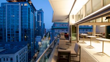 A modern rooftop terrace with seating areas overlooks an urban street with tall buildings at dusk. The interior space is brightly lit.