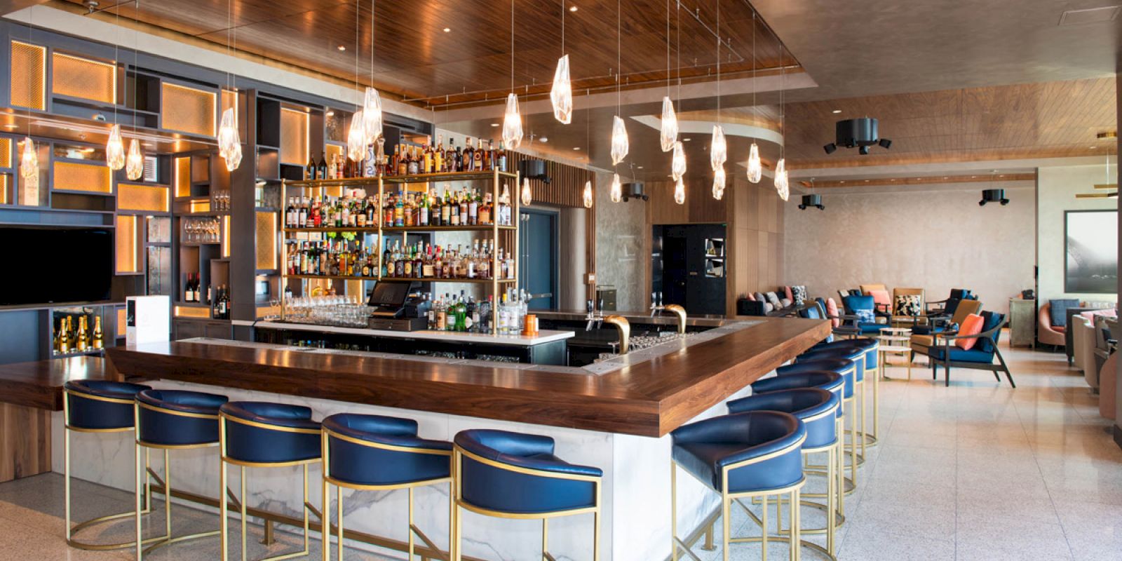 A modern bar with blue cushioned stools and a well-stocked liquor shelf, pendant lights hanging from the ceiling, and a seating area in the background.