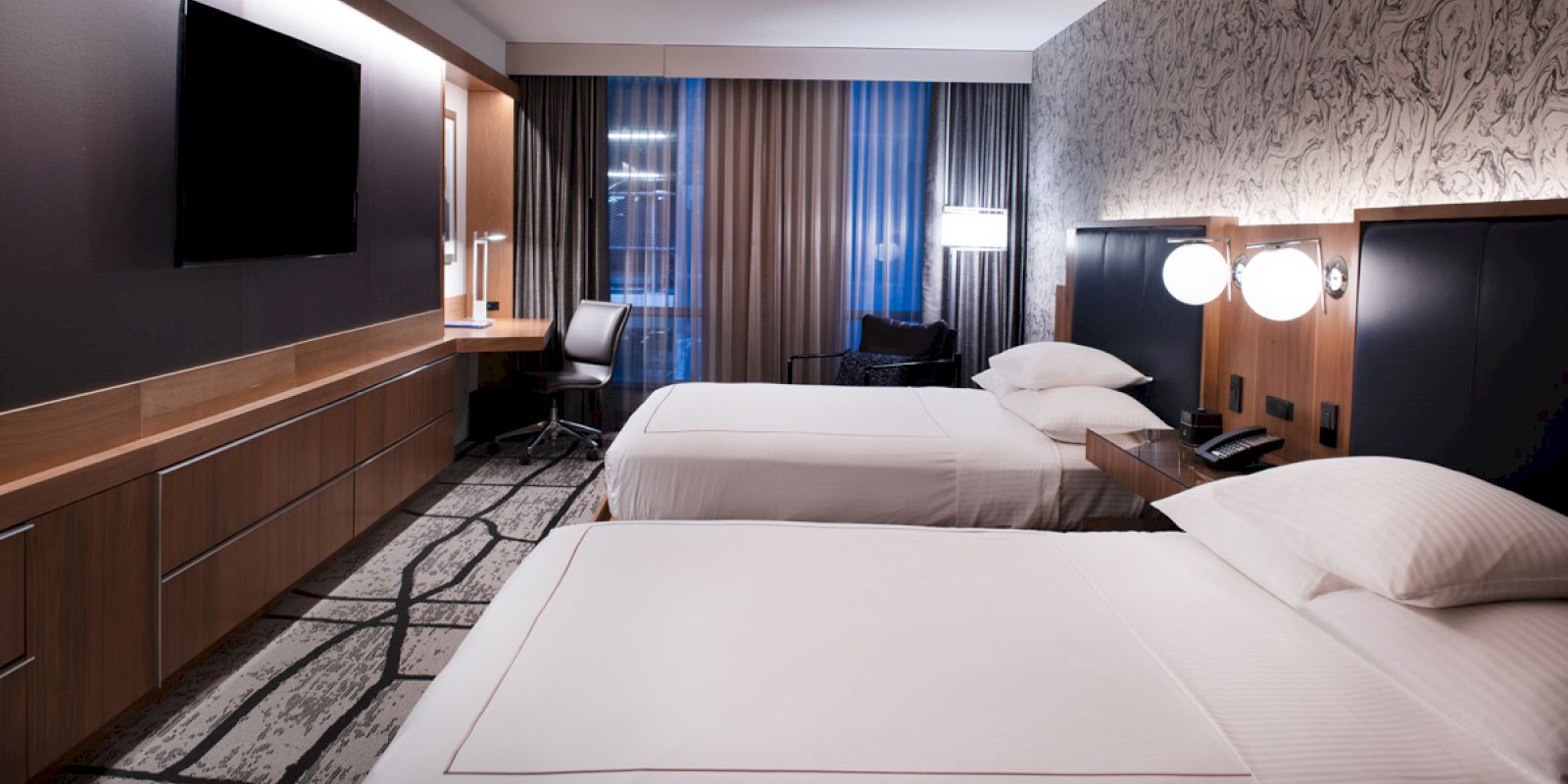 The image shows a modern hotel room with two beds, a flat-screen TV, a desk, and chair, and contemporary decor with ample lighting by the beds.
