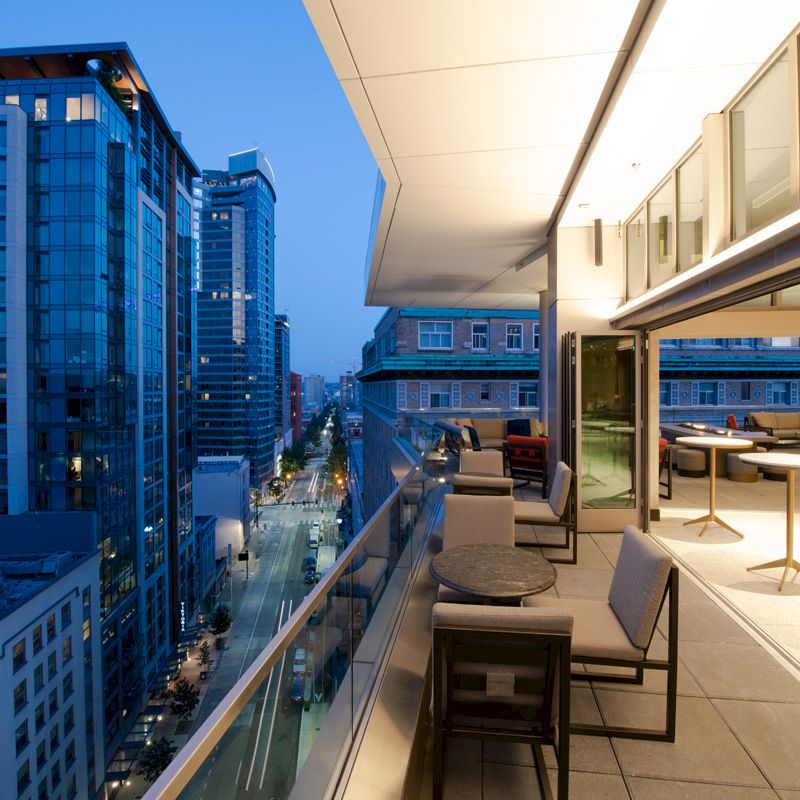 A modern cityscape shows high-rise buildings at dusk, with a stylish balcony featuring tables and chairs extending from a lit indoor space.