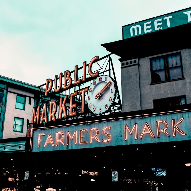 This image shows the famous neon signs of the Public Market and Farmers Market at Pike Place Market in Seattle, with buildings in the background.