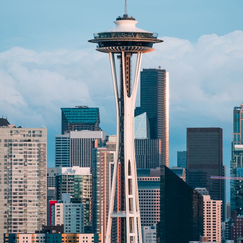 The image shows the Seattle Space Needle with surrounding skyscrapers and buildings in downtown Seattle under a blue sky with some clouds.