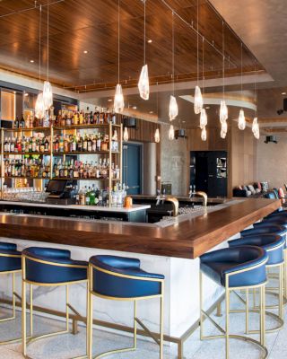 A modern bar with a wooden counter, blue cushioned bar stools, hanging lights, and a fully stocked bar, with additional seating in the background.