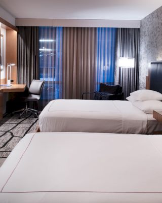 A modern hotel room with two beds, a TV, a desk, and a chair, featuring stylish lighting and decor.
